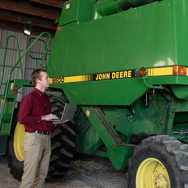 A man with a laptop opened on his palm inspecting a tractor.